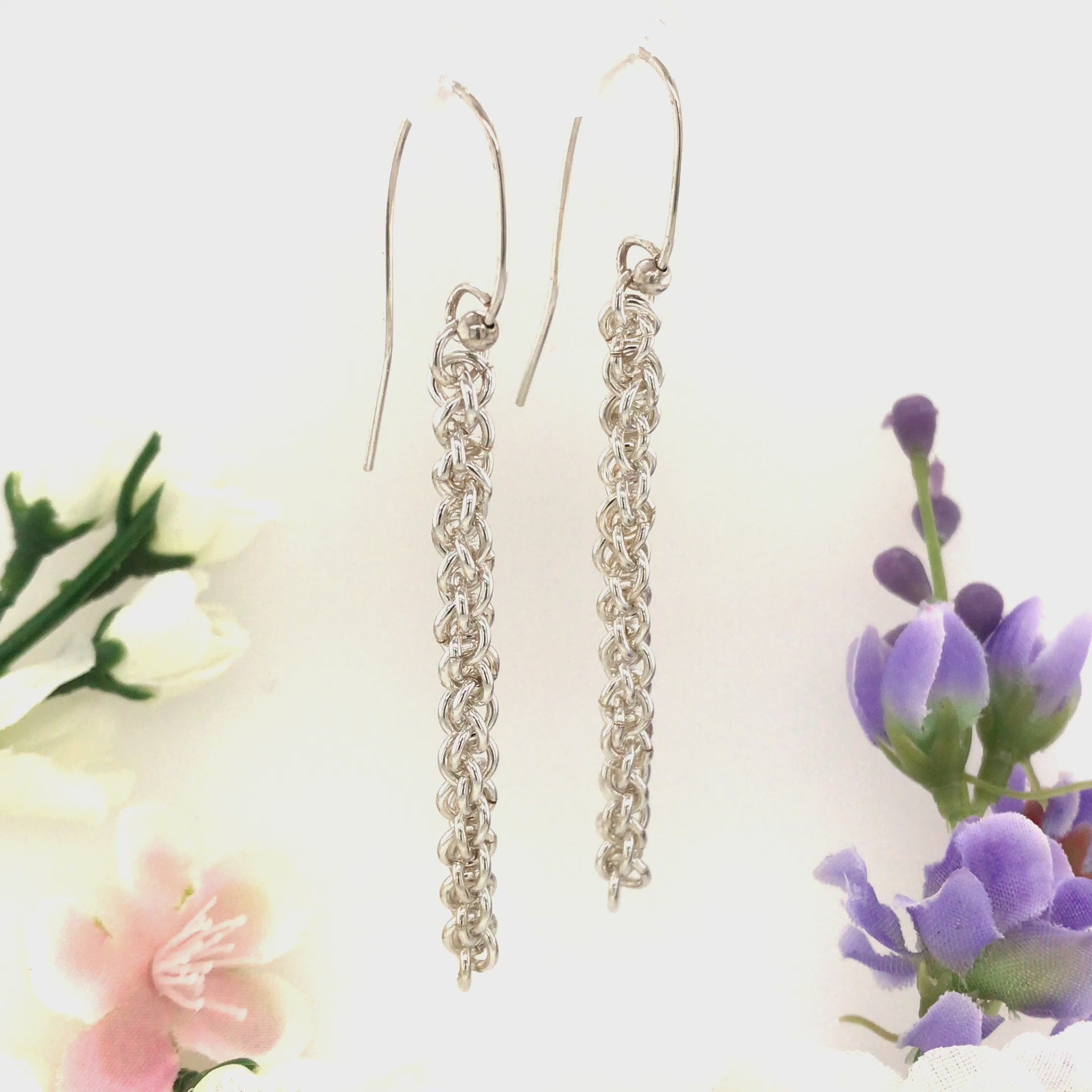 Video, 360 degrees. Bold Spiral Argentium Silver Earrings for Evening (JPL3), swoop ear wire with bead. White, pink, and purple flowers beside earrings.