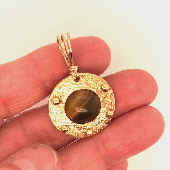 Video, 360 degrees. Tiger's Eye and Brass Circle Pendant being manipulated by hand to show all angles and the shimmer of the tiger's eye.